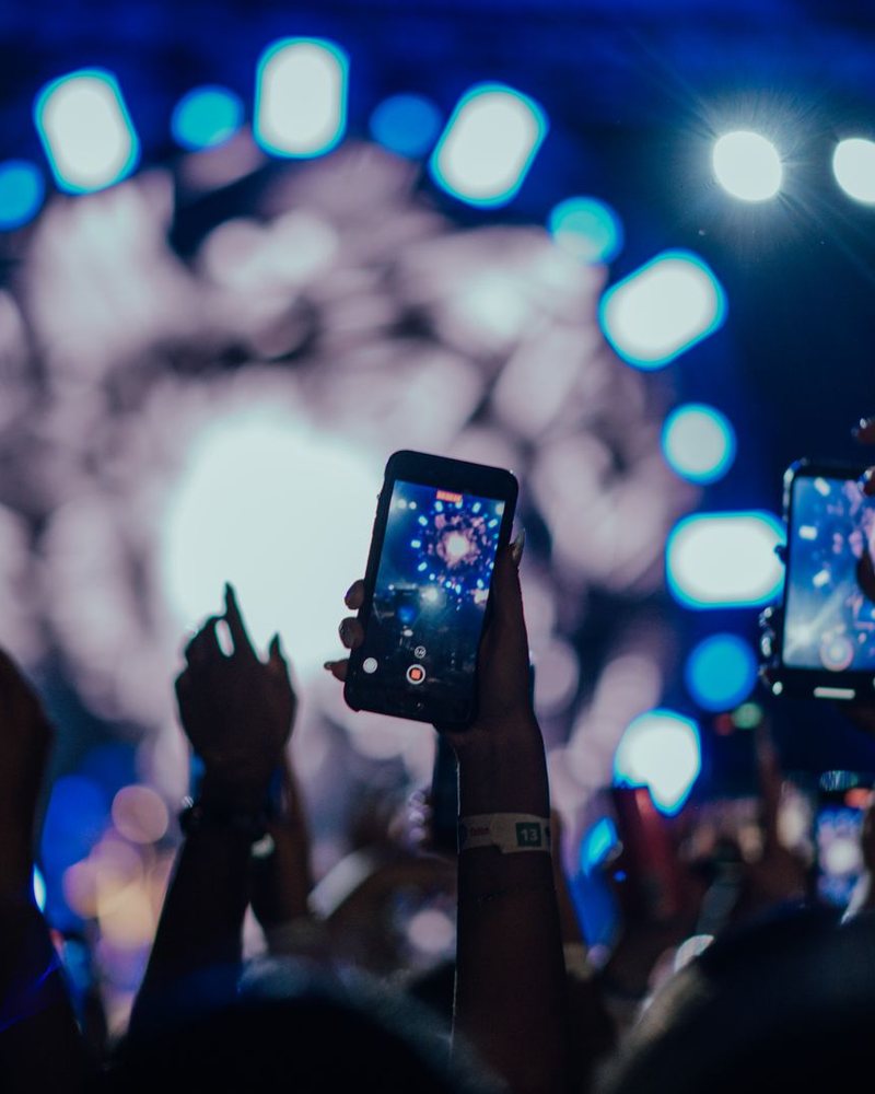 A hand in a concert crowd holds up a phone that films a blue-lit stage; a LIVE mark and platform labels were added to the photo.