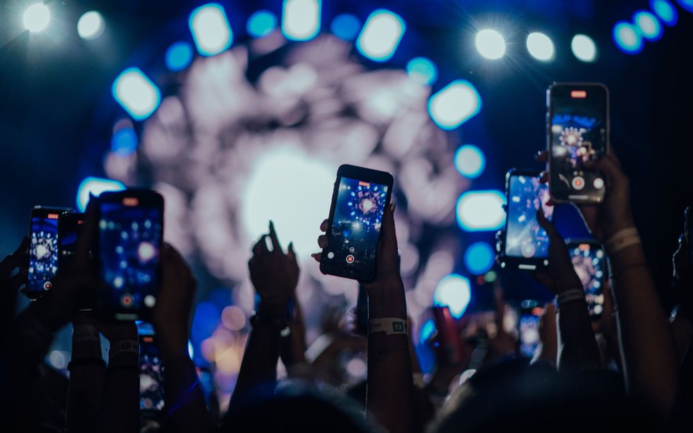 Hands hold up phones to film a concert; a LIVE mark was added.
