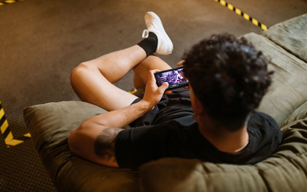 A person lies back on a sofa with their feet up and plays a game on a phone; a LIVE mark was added to the photo.