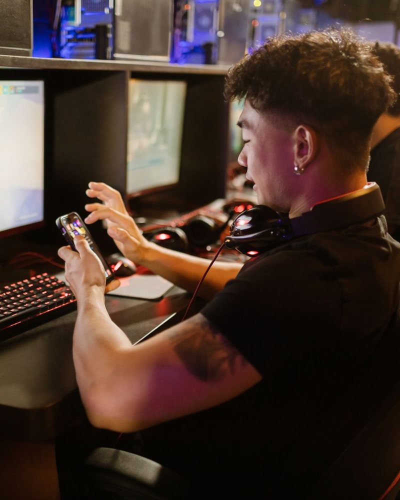 A young man with headphones around his neck holds a phone in front of a monitor and a glowing keyboard; a LIVE mark and platform labels were added to the photo.