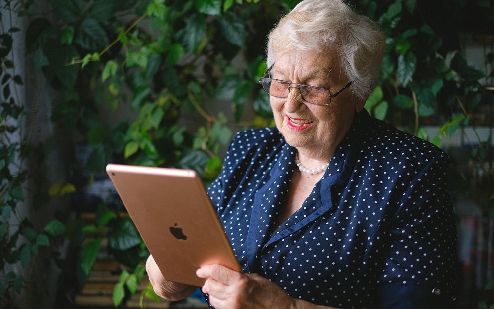 An older woman smiles at a tablet in her hands; a LIVE mark was added to the photo, and the tablet screen is not visible.