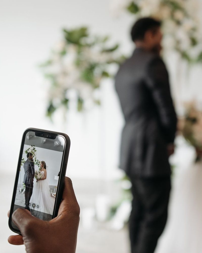 A hand holds a phone that films a bride and groom among white flowers; a LIVE mark and platform labels were added to the photo.